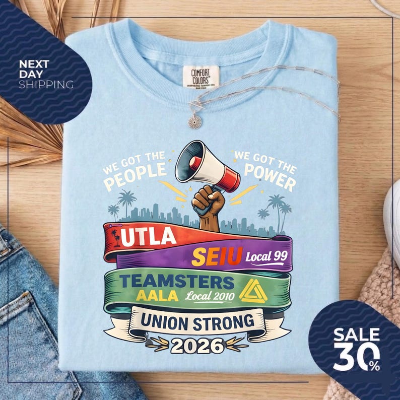 May include: Light blue t-shirt with a graphic featuring a megaphone, a raised fist, and text that reads "We Got The People, We Got The Power, Union Strong 2026." The shirt also displays the logos of UTLA, SEIU, Teamsters, and AALA.