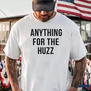 May include: A white short-sleeved t-shirt featuring the black text "ANYTHING FOR THE HUZZ" printed in a bold, sans-serif font across the chest. The shirt is worn with a dark-colored baseball cap, and the fabric has a smooth, clean finish.
