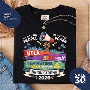 May include: Black t-shirt with the text "WE GOT THE PEOPLE" and "WE GOT THE POWER". The shirt features a megaphone graphic and colorful banners with text including "UTLA", "SEIU", "TEAMSTERS", and "UNION STRONG 2026". A silver necklace is draped on the shirt.