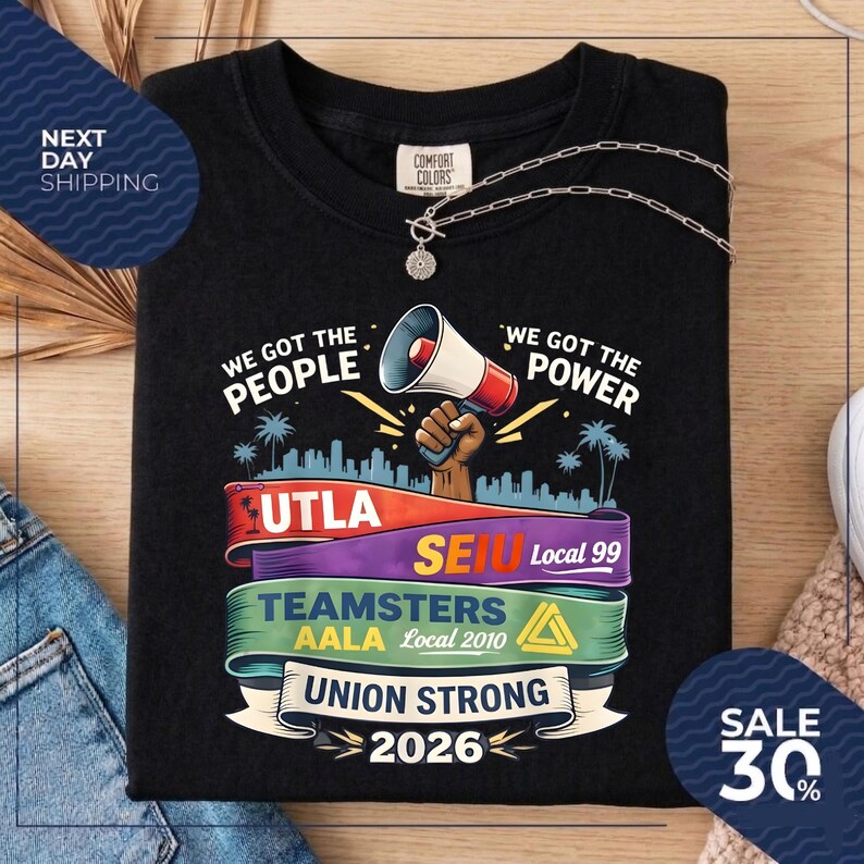 May include: Black t-shirt with the text "WE GOT THE PEOPLE" and "WE GOT THE POWER". The shirt features a megaphone graphic and colorful banners with text including "UTLA", "SEIU", "TEAMSTERS", and "UNION STRONG 2026". A silver necklace is draped on the shirt.