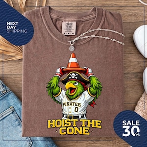K&ouml;nnte beinhalten: Braunes T-Shirt mit einem Cartoon-Papagei, der einen Piratenhut und eine Baseball-Uniform tr&auml;gt und einen orangefarbenen Verkehrskegel h&auml;lt. Das Shirt zeigt "Pirates 0" und "Hoist the Cone". Eine silberne Halskette liegt auf dem Shirt.