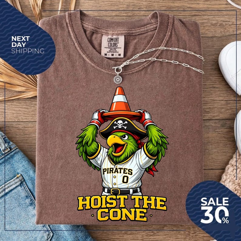 K&ouml;nnte beinhalten: Braunes T-Shirt mit einem Cartoon-Papagei, der einen Piratenhut und eine Baseball-Uniform tr&auml;gt und einen orangefarbenen Verkehrskegel h&auml;lt. Das Shirt zeigt "Pirates 0" und "Hoist the Cone". Eine silberne Halskette liegt auf dem Shirt.