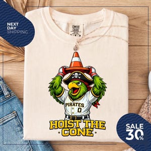 K&ouml;nnte beinhalten: Cremefarbenes T-Shirt mit einem Cartoon-Papagei, der einen Piratenhut und ein Baseball-Trikot mit dem Wort "Pirates" und der Nummer "0" tr&auml;gt. Der Papagei h&auml;lt einen Verkehrskegel. Der Text "HOIST THE CONE" steht darunter.
