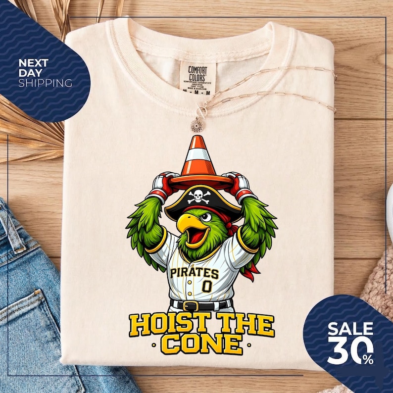 K&ouml;nnte beinhalten: Cremefarbenes T-Shirt mit einem Cartoon-Papagei, der einen Piratenhut und ein Baseball-Trikot mit dem Wort "Pirates" und der Nummer "0" tr&auml;gt. Der Papagei h&auml;lt einen Verkehrskegel. Der Text "HOIST THE CONE" steht darunter.