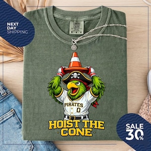 K&ouml;nnte beinhalten: Olivgr&uuml;nes T-Shirt mit einem Cartoon-Papagei, der einen Piratenhut tr&auml;gt und einen orangefarbenen Verkehrskegel h&auml;lt. Das Shirt tr&auml;gt den Text "Pirates 0" und "Hoist the Cone". Eine silberne Halskette ist &uuml;ber dem Shirt drapiert.