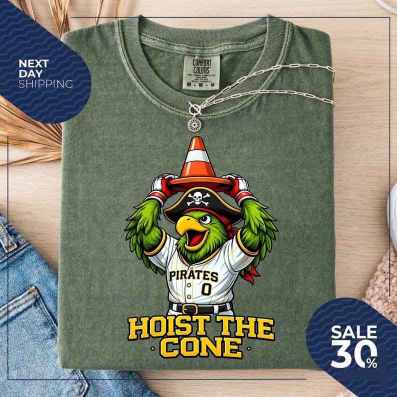 K&ouml;nnte beinhalten: Olivgr&uuml;nes T-Shirt mit einem Cartoon-Papagei, der einen Piratenhut tr&auml;gt und einen orangefarbenen Verkehrskegel h&auml;lt. Das Shirt tr&auml;gt den Text "Pirates 0" und "Hoist the Cone". Eine silberne Halskette ist &uuml;ber dem Shirt drapiert.