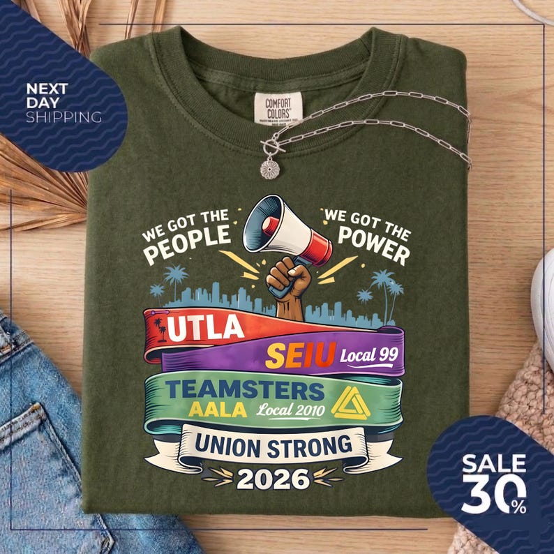 May include: Olive green t-shirt with the phrases "We Got The People" and "We Got The Power". The shirt features a megaphone graphic and text for UTLA, SEIU, Teamsters, AALA, and "Union Strong 2026". A silver necklace is draped across the shirt.