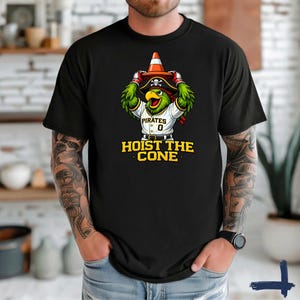 K&ouml;nnte beinhalten: Schwarzes T-Shirt mit einem Cartoon-Papagei, der einen Piratenhut tr&auml;gt und einen Verkehrskegel h&auml;lt. Der Papagei tr&auml;gt ein Pirates-Baseball-Trikot mit der Nummer 0. Der Text "HOIST THE CONE" ist darunter in Gelb gedruckt.
