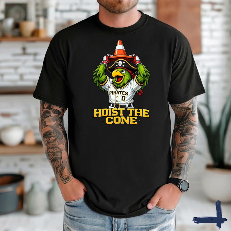 K&ouml;nnte beinhalten: Schwarzes T-Shirt mit einem Cartoon-Papagei, der einen Piratenhut tr&auml;gt und einen Verkehrskegel h&auml;lt. Der Papagei tr&auml;gt ein Pirates-Baseball-Trikot mit der Nummer 0. Der Text "HOIST THE CONE" ist darunter in Gelb gedruckt.