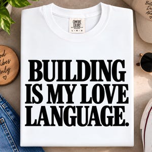 Pode incluir: Uma t-shirt branca de manga curta com o texto preto "BUILDING IS MY LOVE LANGUAGE." impresso numa fonte serifada em negrito no centro. A t-shirt tem um decote redondo e uma etiqueta da marca Comfort Colors visível na gola interior.