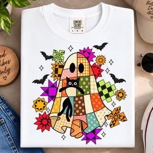 Puede incluir: Una camiseta blanca con un gráfico de un fantasma cubierto con un patrón de colcha de retazos colorida, sosteniendo un gato negro. El diseño incluye varias formas de estrellas geométricas en colores brillantes y murciélagos negros.
