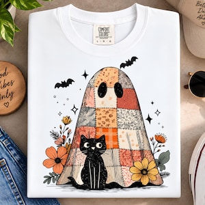 Może przedstawiać: Biały t-shirt z grafiką ducha przykrytego patchworkową kołdrą w odcieniach pomarańczu i brązu. Przed duchem siedzi czarny kot, otoczony pomarańczowymi i żółtymi kwiatami, małymi czarnymi nietoperzami oraz ikonami gwiazdek.