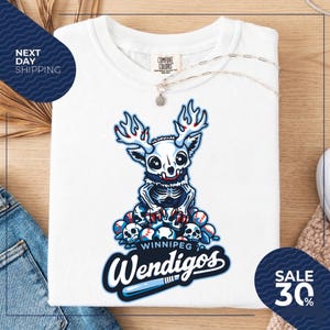 Puede incluir: Camiseta blanca con un diseño de Wendigo de dibujos animados, con un esqueleto, cornamentas y pelotas de béisbol. El texto "Winnipeg Wendigos" se muestra en una fuente estilizada. Un collar está drapeado sobre la camiseta.
