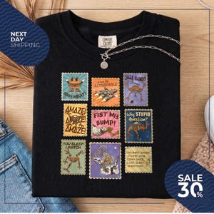 May include: Black t-shirt with a collage of colorful, vintage-style postage stamp designs. The stamps feature various illustrations and phrases, including "100% MEAT!", "JAZZ HANDS", and "FIST MY BUMP!" A silver necklace with a pendant is draped across the shirt.