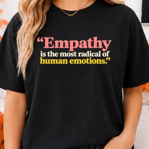 Op de afbeelding: Een zwart t-shirt met korte mouwen en de tekst "Empathy is the most radical of human emotions." Het woord Empathy is in het roze gedrukt, terwijl de rest van de tekst in het geel is gedrukt, gecentreerd op de voorzijde van het kledingstuk.