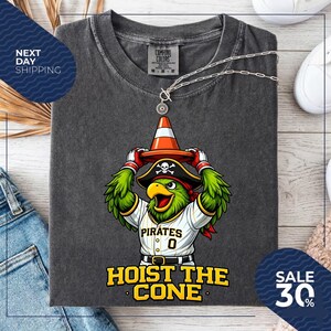 K&ouml;nnte beinhalten: Dunkelgraues T-Shirt mit einem Cartoon-Papagei, der einen Piratenhut tr&auml;gt und einen orangefarbenen Verkehrskegel h&auml;lt. Das Shirt tr&auml;gt den Text "HOIST THE CONE" und "PIRATES 0". Eine silberne Halskette ist &uuml;ber dem Shirt drapiert.