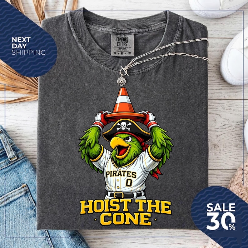 K&ouml;nnte beinhalten: Dunkelgraues T-Shirt mit einem Cartoon-Papagei, der einen Piratenhut tr&auml;gt und einen orangefarbenen Verkehrskegel h&auml;lt. Das Shirt tr&auml;gt den Text "HOIST THE CONE" und "PIRATES 0". Eine silberne Halskette ist &uuml;ber dem Shirt drapiert.
