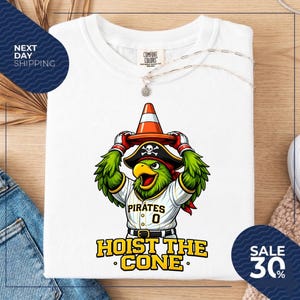 K&ouml;nnte beinhalten: Wei&szlig;es T-Shirt mit einem Cartoon-Papagei, der einen Piratenhut tr&auml;gt und einen orangefarbenen Verkehrskegel h&auml;lt. Der Papagei tr&auml;gt ein Pirates-Baseballtrikot mit der Nummer 0. Der Text "HOIST THE CONE" steht darunter.