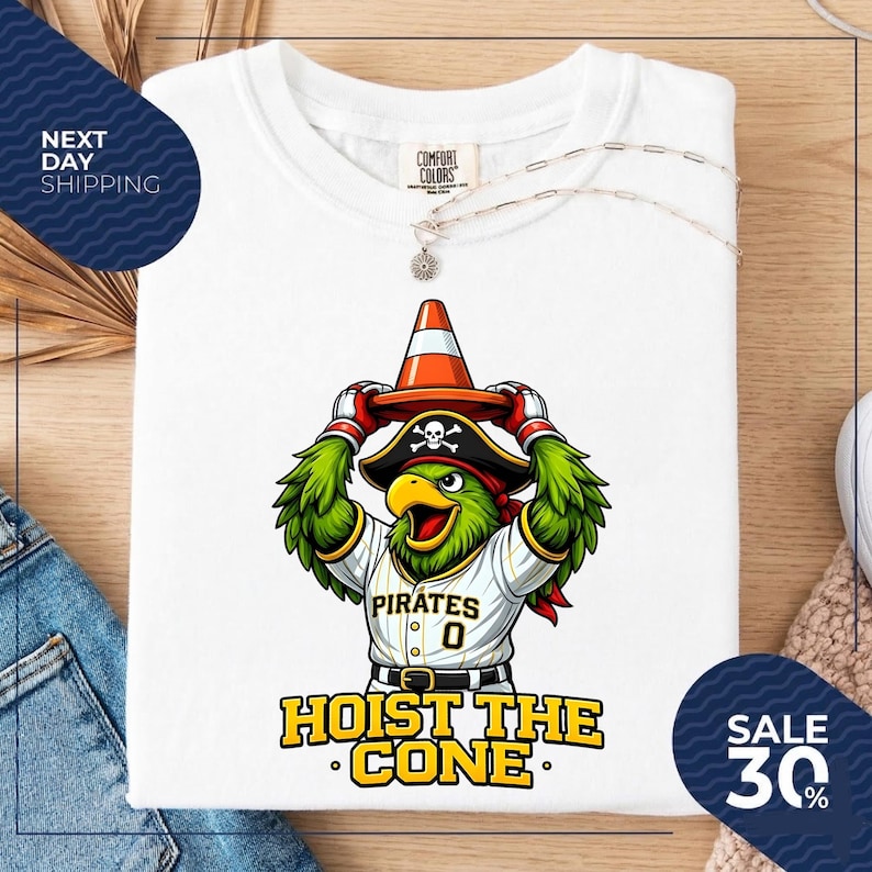 K&ouml;nnte beinhalten: Wei&szlig;es T-Shirt mit einem Cartoon-Papagei, der einen Piratenhut tr&auml;gt und einen orangefarbenen Verkehrskegel h&auml;lt. Der Papagei tr&auml;gt ein Pirates-Baseballtrikot mit der Nummer 0. Der Text "HOIST THE CONE" steht darunter.