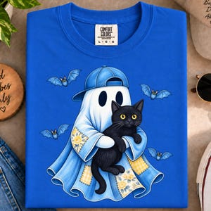 Puede incluir: Una camiseta azul brillante de cuello redondo con un gráfico de un fantasma que lleva una gorra de béisbol azul y sostiene un gato negro. El fantasma está envuelto en una tela azul con parches florales y de cuadros amarillos, rodeado de cuatro pequeños murciélagos azules.