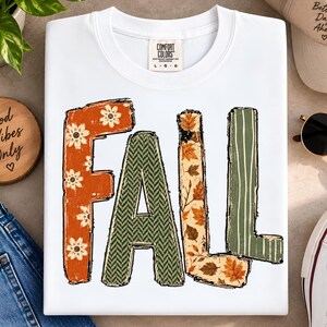 Könnte beinhalten: Ein weißes T-Shirt mit Rundhalsausschnitt und dem Wort FALL in großen Blockbuchstaben. Das F ist orange mit weißen Blumen, das A ist grün mit Fischgrätenmuster, das L ist beige mit Herbstblättern und das zweite L ist grün mit weißen Streifen.