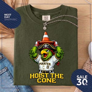 K&ouml;nnte beinhalten: Olivgr&uuml;nes T-Shirt mit einem Cartoon-Papagei, der einen Piratenhut tr&auml;gt und einen Verkehrskegel h&auml;lt. Das Shirt hat den Text "Pirates 0" und "Hoist the Cone". Eine silberne Halskette ist &uuml;ber dem Shirt drapiert.