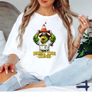 K&ouml;nnte beinhalten: Wei&szlig;es T-Shirt mit einem Cartoon-Papagei, der einen Piratenhut tr&auml;gt und einen Verkehrskegel h&auml;lt. Der Papagei tr&auml;gt ein Baseball-Trikot mit dem Wort "Pirates" und der Nummer "0". Der Text "HOIST THE CONE" ist darunter gedruckt.