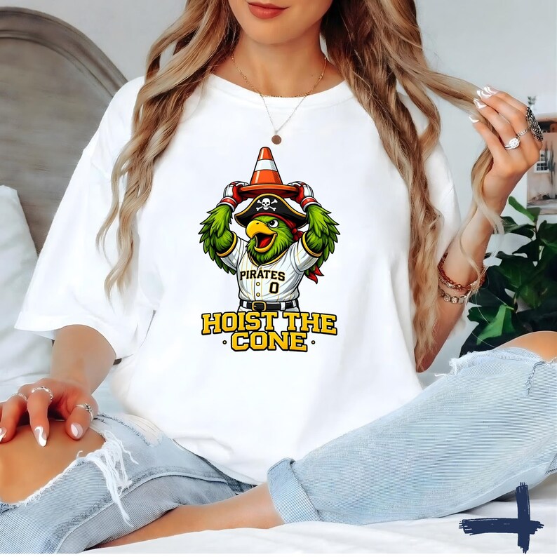 K&ouml;nnte beinhalten: Wei&szlig;es T-Shirt mit einem Cartoon-Papagei, der einen Piratenhut tr&auml;gt und einen Verkehrskegel h&auml;lt. Der Papagei tr&auml;gt ein Baseball-Trikot mit dem Wort "Pirates" und der Nummer "0". Der Text "HOIST THE CONE" ist darunter gedruckt.