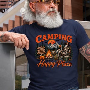 May include: A navy blue t-shirt featuring a graphic of a skeleton sitting in a camping chair by a campfire. The design includes mountains, pine trees, a pumpkin, and text that reads CAMPING is my Happy Place, with signs for CAMPFIRE, S'MORES, and SPOOKY VIBES.