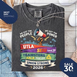 May include: Dark gray t-shirt with the text "WE GOT THE PEOPLE" and "WE GOT THE POWER". The shirt features a graphic of a megaphone and the words "UNION STRONG 2026". A silver necklace is draped across the shirt.