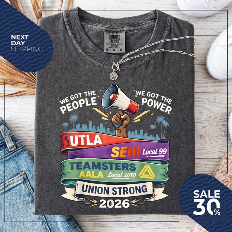 May include: Dark gray t-shirt with the text "WE GOT THE PEOPLE" and "WE GOT THE POWER". The shirt features a graphic of a megaphone and the words "UNION STRONG 2026". A silver necklace is draped across the shirt.