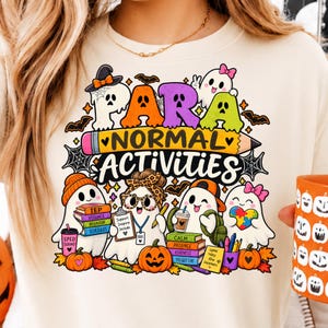 Può includere: Una felpa color crema con una grafica colorata che riporta il testo "PARA NORMAL ACTIVITIES". Il design include fantasmi dei cartoni animati, zucche, libri etichettati "IEP", "VISUALS", "BEHAVIOR", "SCHEDULES", "CALM", "PATIENCE", "KINDNESS" e un cuore puzzle.