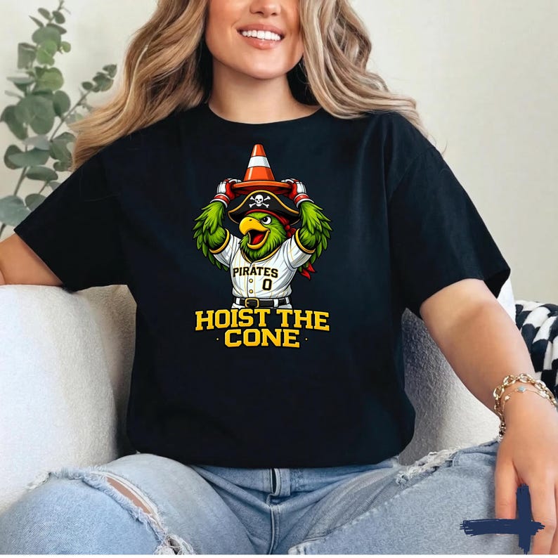 K&ouml;nnte beinhalten: Schwarzes T-Shirt mit einem Cartoon-Papagei, der einen Piratenhut tr&auml;gt und einen Verkehrskegel h&auml;lt. Der Papagei tr&auml;gt ein Baseball-Trikot mit der Aufschrift "Pirates" und "0". Der Text "HOIST THE CONE" ist gelb gedruckt.