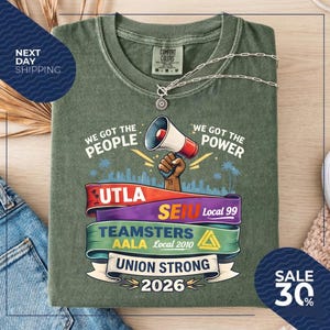 May include: Olive green t-shirt with the phrases "We Got The People" and "We Got The Power" printed on it. The shirt features a megaphone graphic and several union logos. A silver necklace is draped across the shirt.