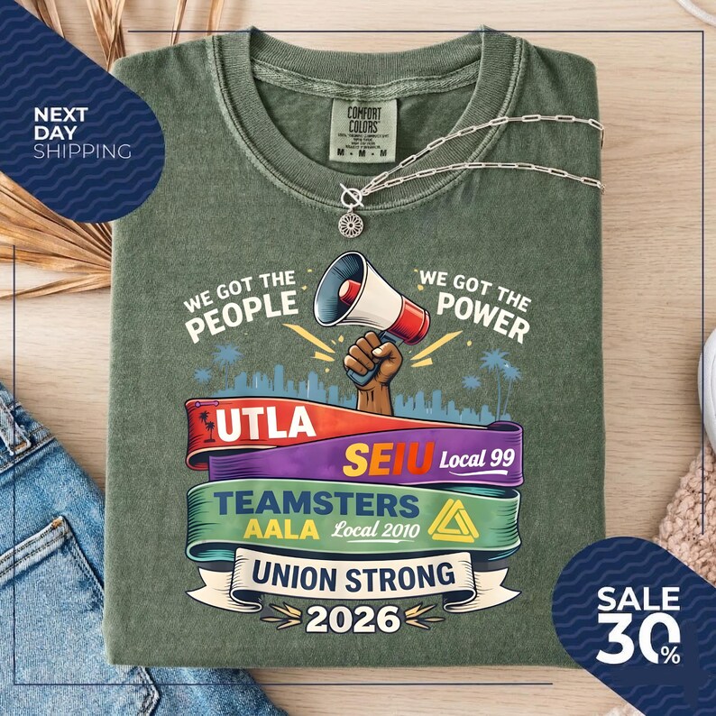 May include: Olive green t-shirt with the phrases "We Got The People" and "We Got The Power" printed on it. The shirt features a megaphone graphic and several union logos. A silver necklace is draped across the shirt.