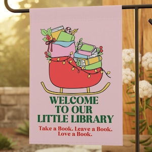 May include: A pink garden flag with the text "WELCOME TO OUR LITTLE LIBRARY" and the phrase "Take a Book. Leave a Book. Love a Book." A red sleigh filled with books and Christmas lights is depicted above the text.