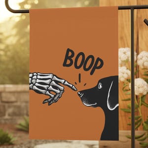 May include: Orange garden flag with a black dog graphic and the word "BOOP". A skeleton hand is touching the dog's nose. The flag is displayed on a black metal stand in a garden setting.