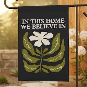 May include: Black garden flag with the text "IN THIS HOME WE BELIEVE IN" and a white flower design. The flag also includes the words "inclusion", "science", "love is love", "women's rights", "human rights", and "diversity".
