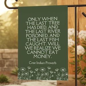 May include: A green garden flag with white text and a white floral border. The text reads: 'ONLY WHEN THE LAST TREE HAS DIED, AND THE LAST RIVER POISONED, AND THE LAST FISH CAUGHT, WILL WE REALIZE WE CANNOT EAT MONEY. Cree Indian Proverb'