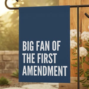 Puede incluir: Una bandera de jardín azul marino con el texto blanco "BIG FAN OF THE FIRST AMENDMENT". La bandera está en un soporte de metal negro en un jardín con adoquines y flores blancas.