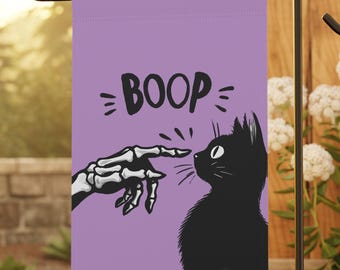 May include: A purple garden flag featuring a black cat and a skeleton hand touching noses. The word "BOOP" is above the cat. The flag is displayed on a black metal stand in a garden setting.