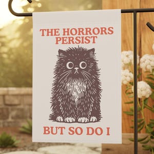 Può includere: Una bandiera da giardino su un supporto in metallo nero. La bandiera presenta un'illustrazione di un gatto dei cartoni animati in marrone e bianco, con il testo "THE HORRORS PERSIST BUT SO DO I" in rosso. La bandiera è in un giardino.
