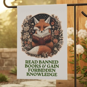 May include: A white garden flag with a fox reading a book, encircled by flowers and leaves. The flag displays the text "READ BANNED BOOKS & GAIN FORBIDDEN KNOWLEDGE" in green. The flag is mounted on a black metal garden stake.