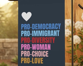 Puede incluir: Una bandera de jardín azul marino con un corazón blanco y las palabras "PRO-DEMOCRACY, PRO-IMMIGRANT, PRO-DIVERSITY, PRO-WOMAN, PRO-CHOICE, PRO-LOVE" en varios colores. La bandera está en un soporte de metal negro en un jardín.