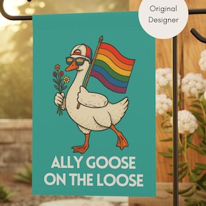 May include: A teal garden flag features a cartoon goose wearing sunglasses and a hat, holding a rainbow flag and flowers. The text "ALLY GOOSE ON THE LOOSE" is displayed below the goose. The flag is mounted on a black metal stand.