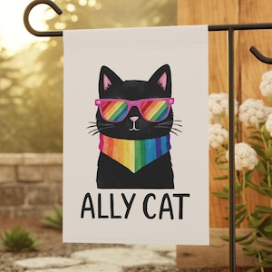 May include: A garden flag featuring a black cat wearing pink sunglasses and a rainbow scarf. The flag reads "ALLY CAT" in black text. The flag is displayed on a black metal stand in a garden setting.