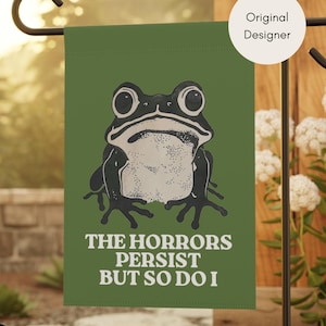 May include: A rectangular olive green garden flag featuring a black and white illustration of a frog with large eyes. Below the frog, the text reads THE HORRORS PERSIST BUT SO DO I in white capital letters. A circular badge says Original Designer.