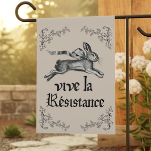 May include: A garden flag featuring a running rabbit with a bow, surrounded by floral accents. The flag has the text "vive la Résistance" in black gothic lettering. The flag is displayed on a black metal stand in a garden setting.