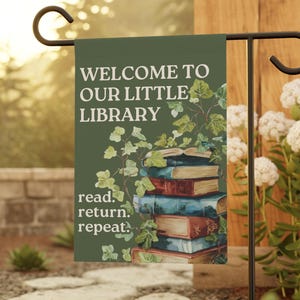 Puede incluir: Una bandera de jardín verde con el texto "WELCOME TO OUR LITTLE LIBRARY" y "read. return. repeat." Una pila de libros con hiedra está ilustrada debajo del texto. La bandera se muestra en un soporte de metal negro en un jardín.