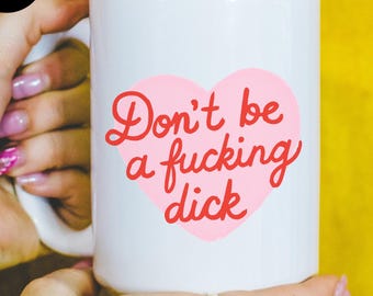 Może przedstawiać: Biały ceramiczny kubek z różowym sercem i napisem "Don't be a fucking dick" w czerwonym skrypcie. Kubek trzymany jest przez osobę z tatuażami i złotymi pierścionkami na żółtym tle.